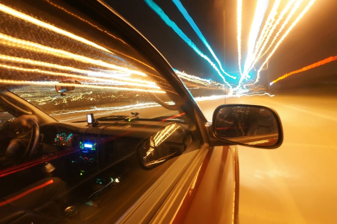 Glowing orange and blue light trails stretch across a dark car dashboard view during a fast night drive.