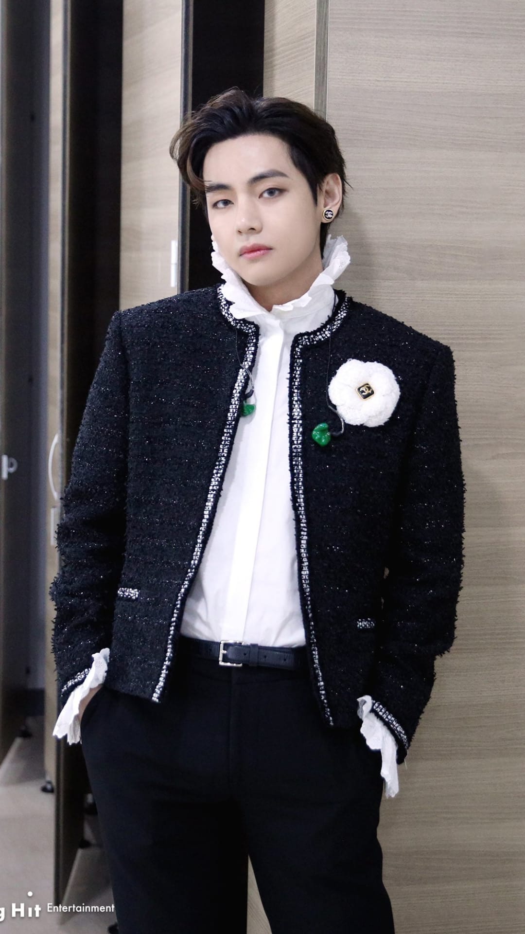 Kim Taehyung wearing a black tweed jacket with a white floral brooch and a ruffled white collar shirt.