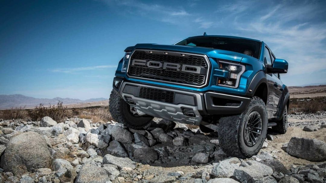 Blue Ford Raptor pickup truck driving over large rocks in a desert with mountains and a clear blue sky.