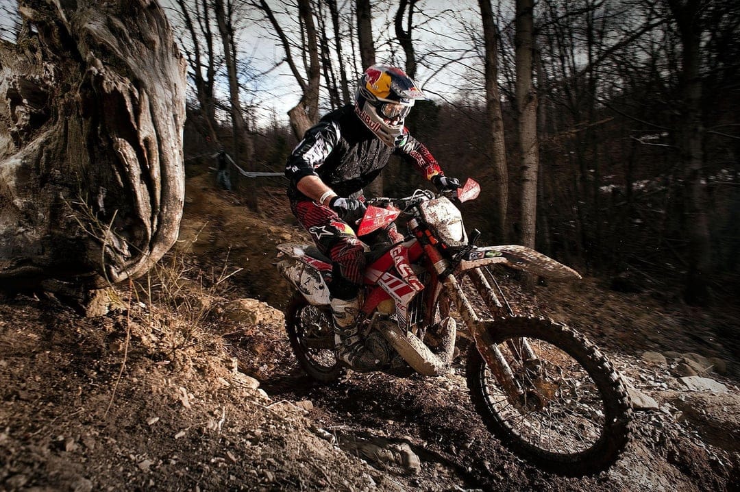 Dirt bike rider on a red motorcycle navigates a muddy forest trail with sunlight filtering through bare trees.