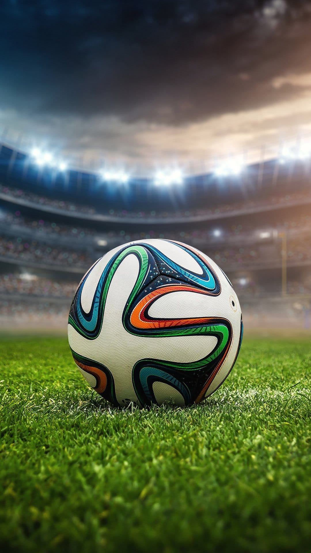 Soccer ball resting on stadium grass with blurred floodlights and a dramatic cloudy sky in the background.