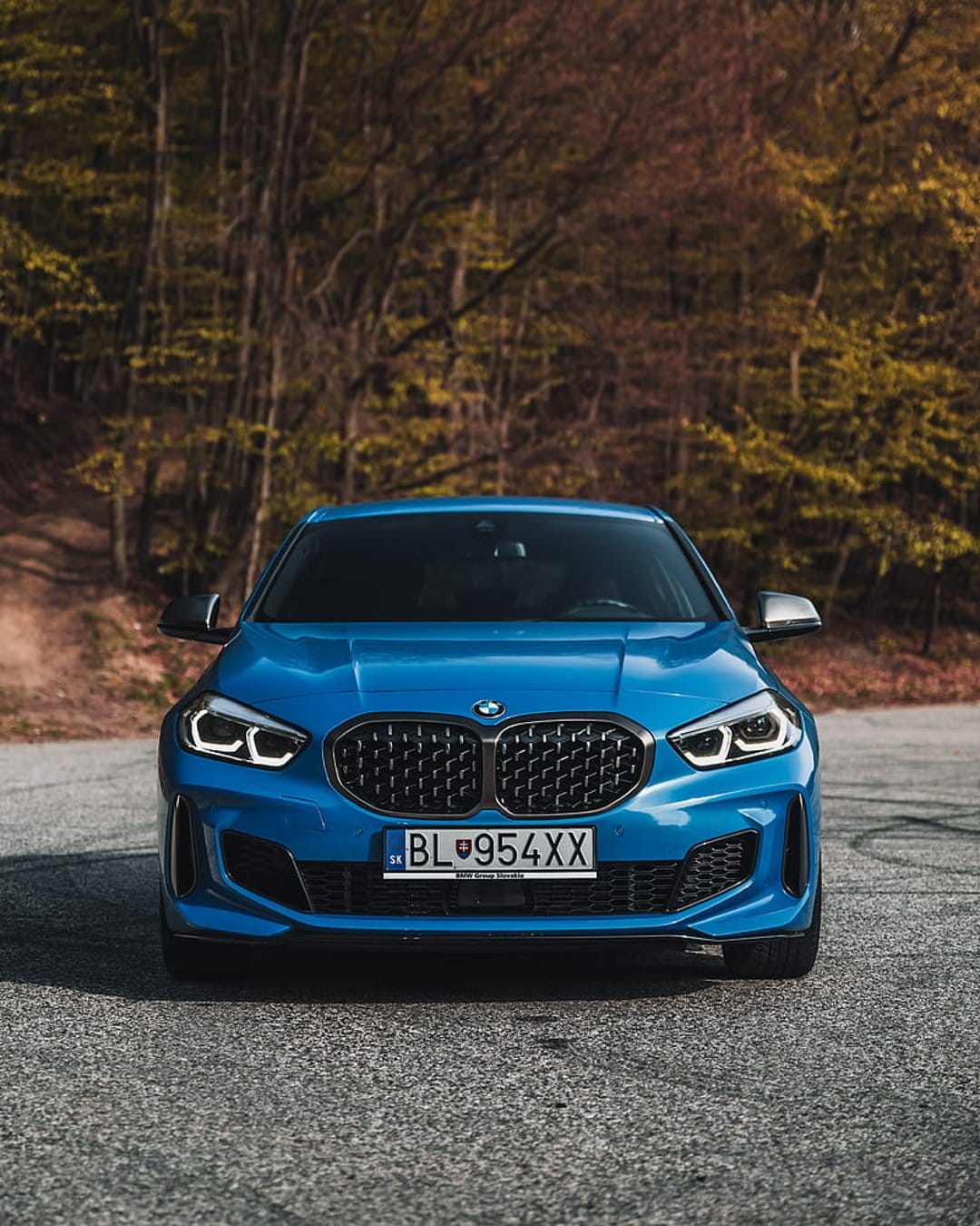 Blue BMW M135i with LED headlights parked on a paved forest road during autumn with blurred trees behind it.