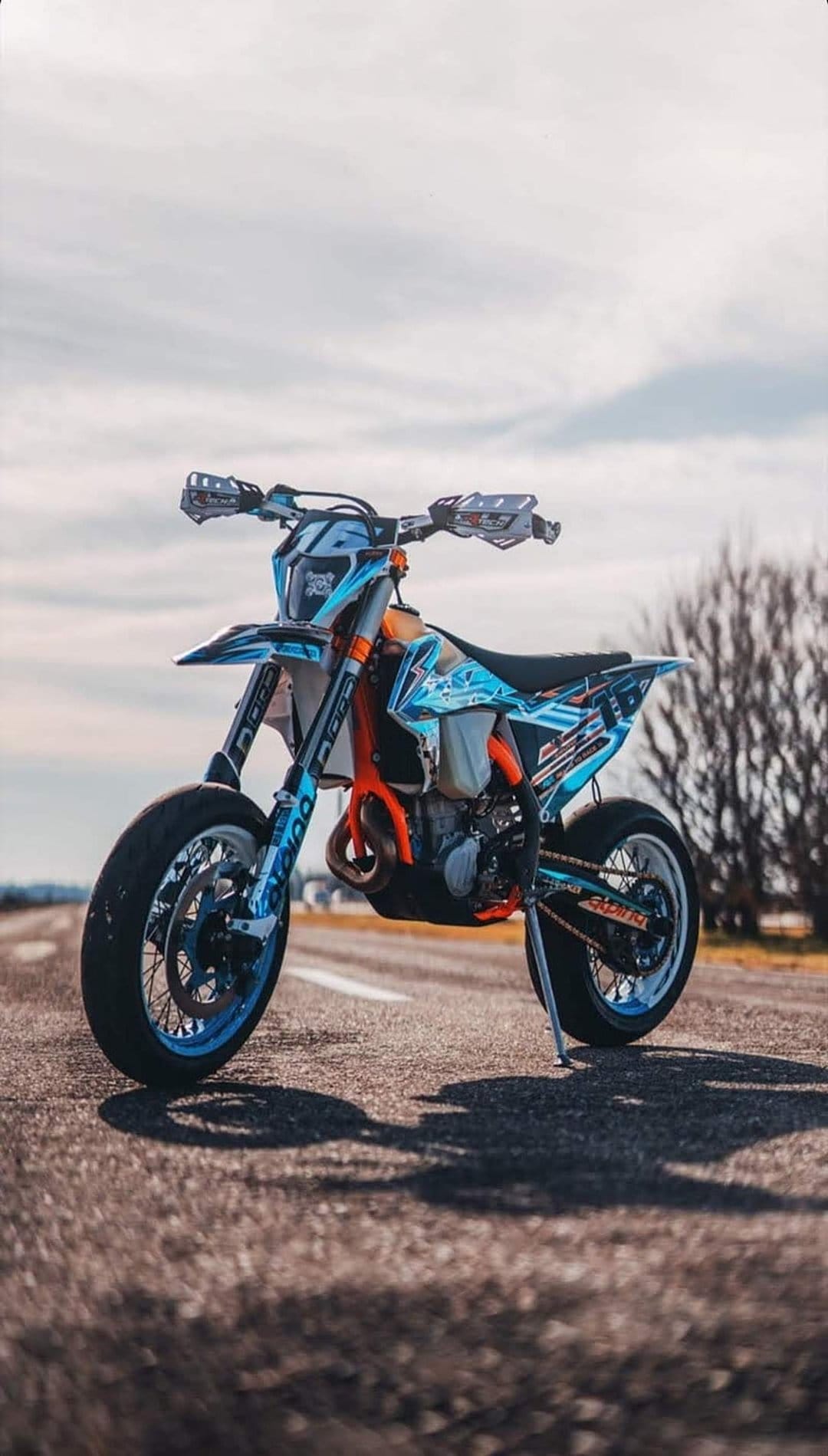Supermoto motorcycle with blue and orange graphics parked on asphalt featuring performance tires and handguards.