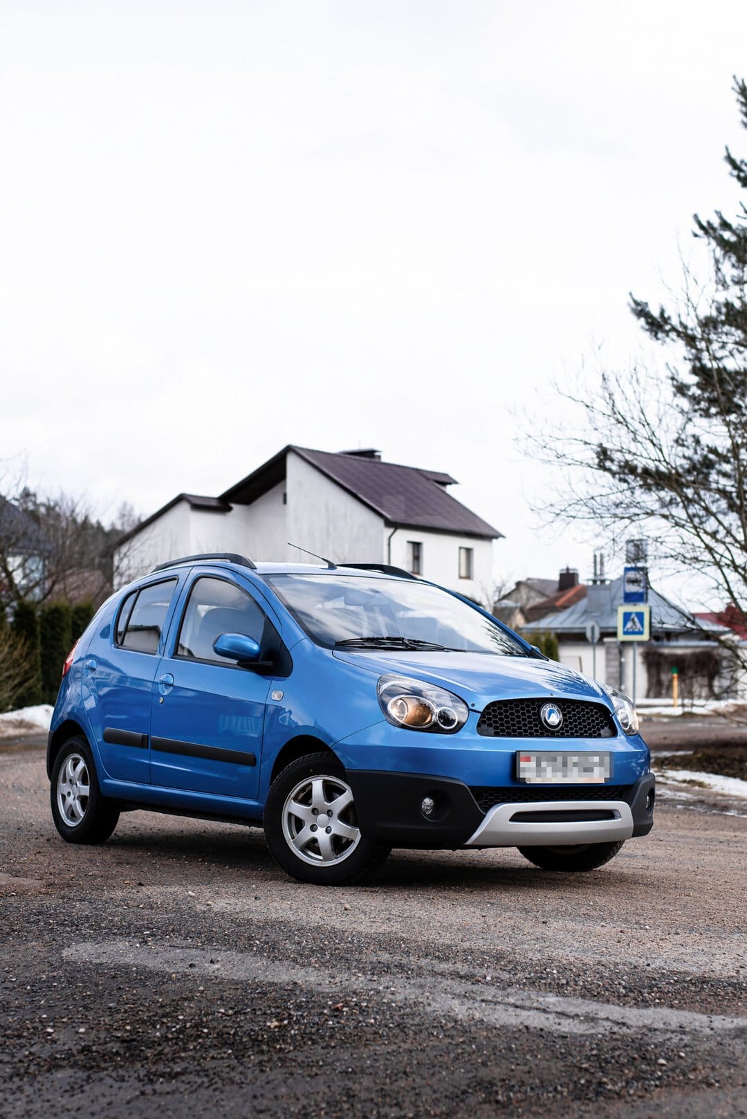 Blue Geely Panda GX2 micro-SUV on a gravel road against a white house and overcast sky in portrait view.