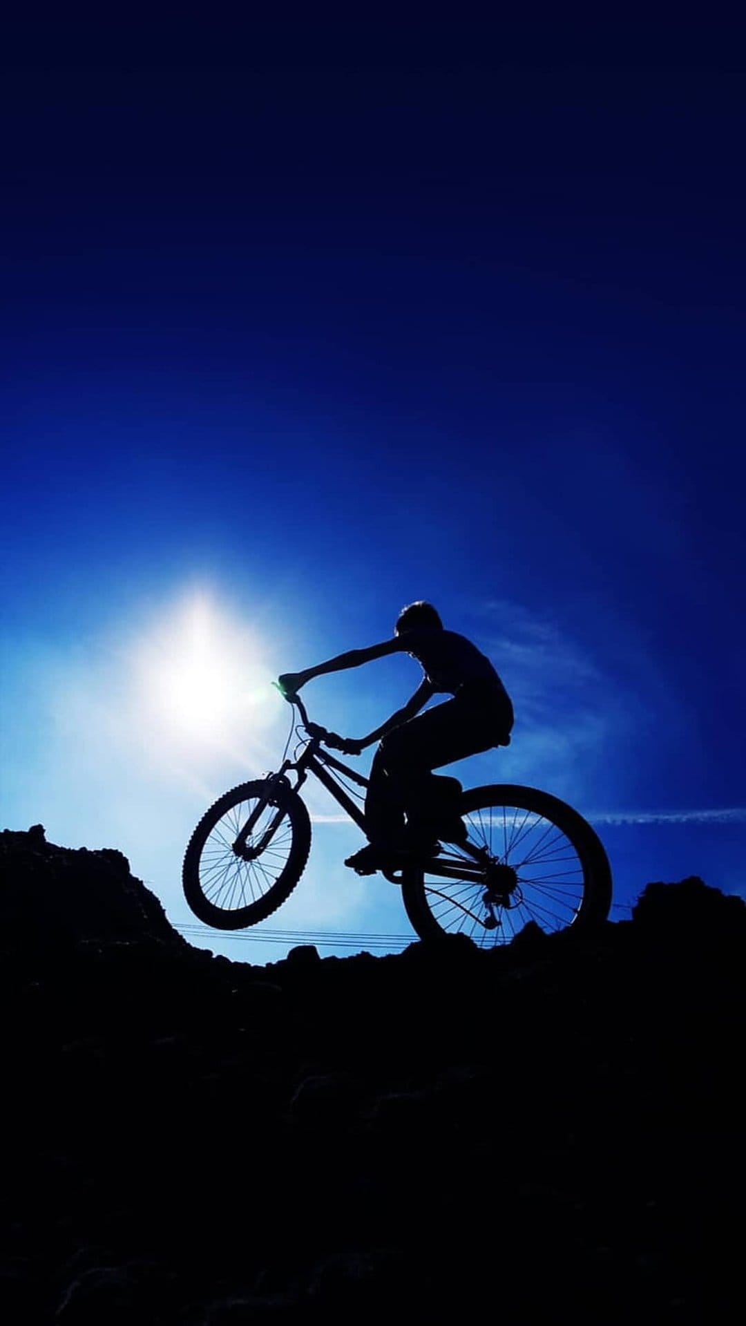 Mountain biker silhouette jumping in mid-air with a bright sun flare against a deep blue sky over dark rocks.