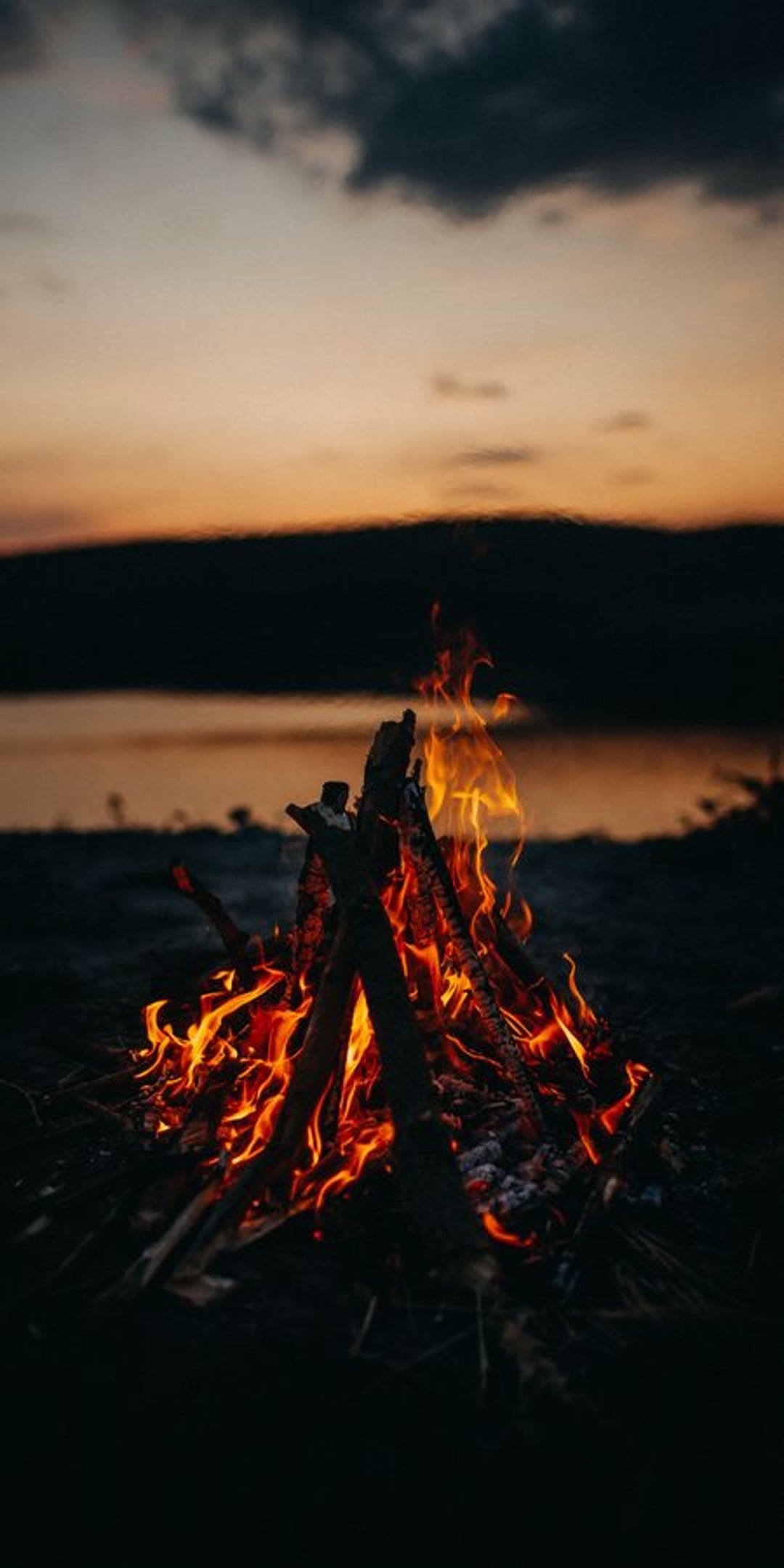 Crackling orange bonfire burns on a rocky shore with light reflecting on the water at dusk under a purple sky.