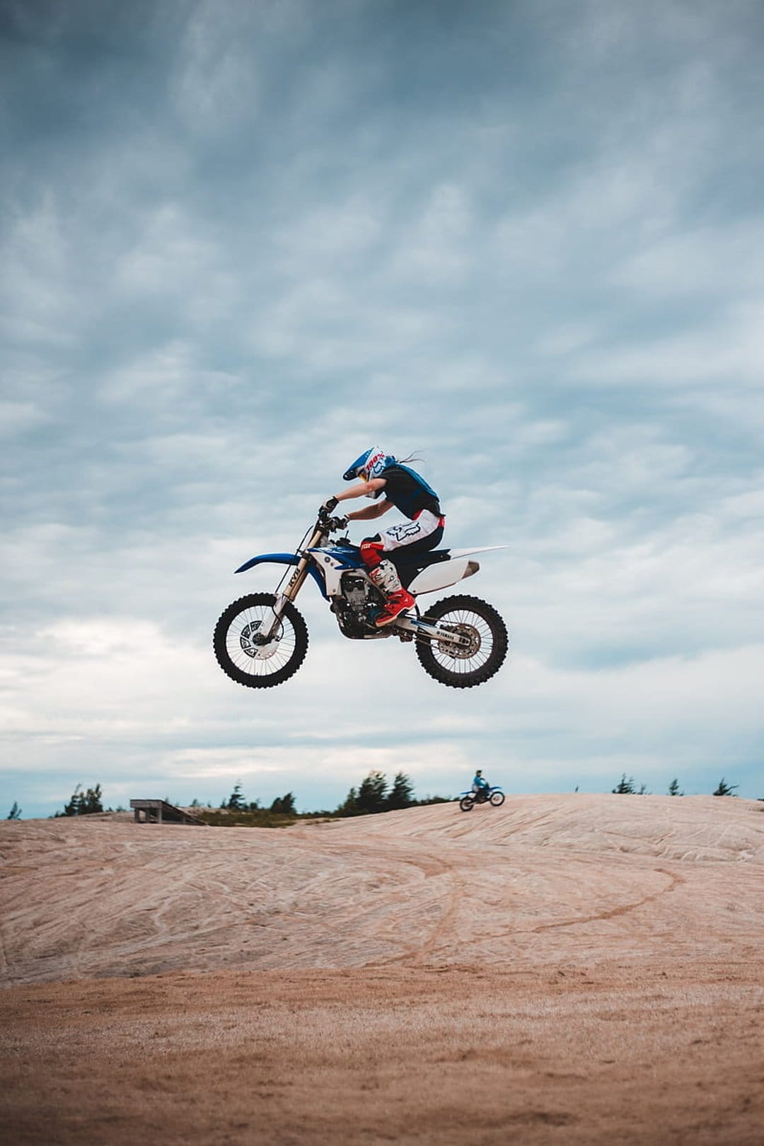 Motocross rider in gear jumps a dirt bike high into a cloudy sky above a sandy racing track and distant trees.
