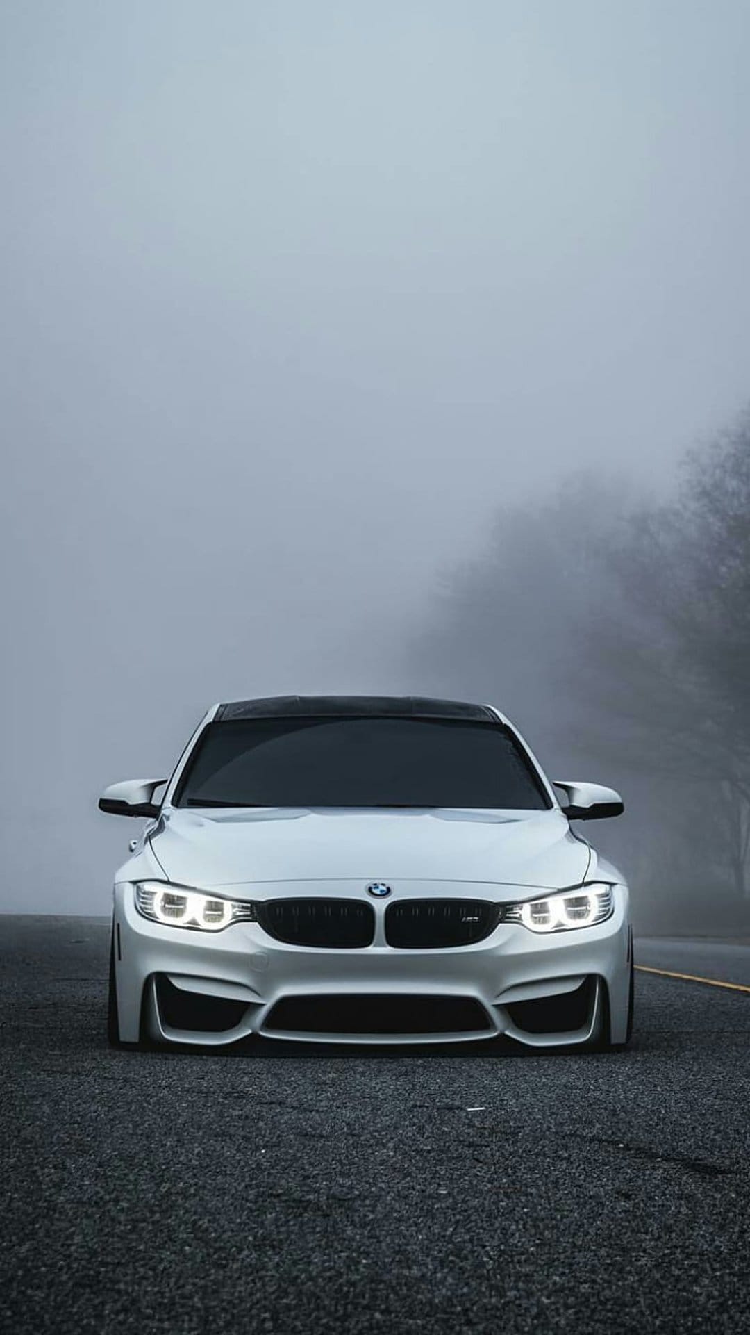 White BMW M4 coupe with lit headlights on a dark road surrounded by heavy fog and blurred trees in the distance.