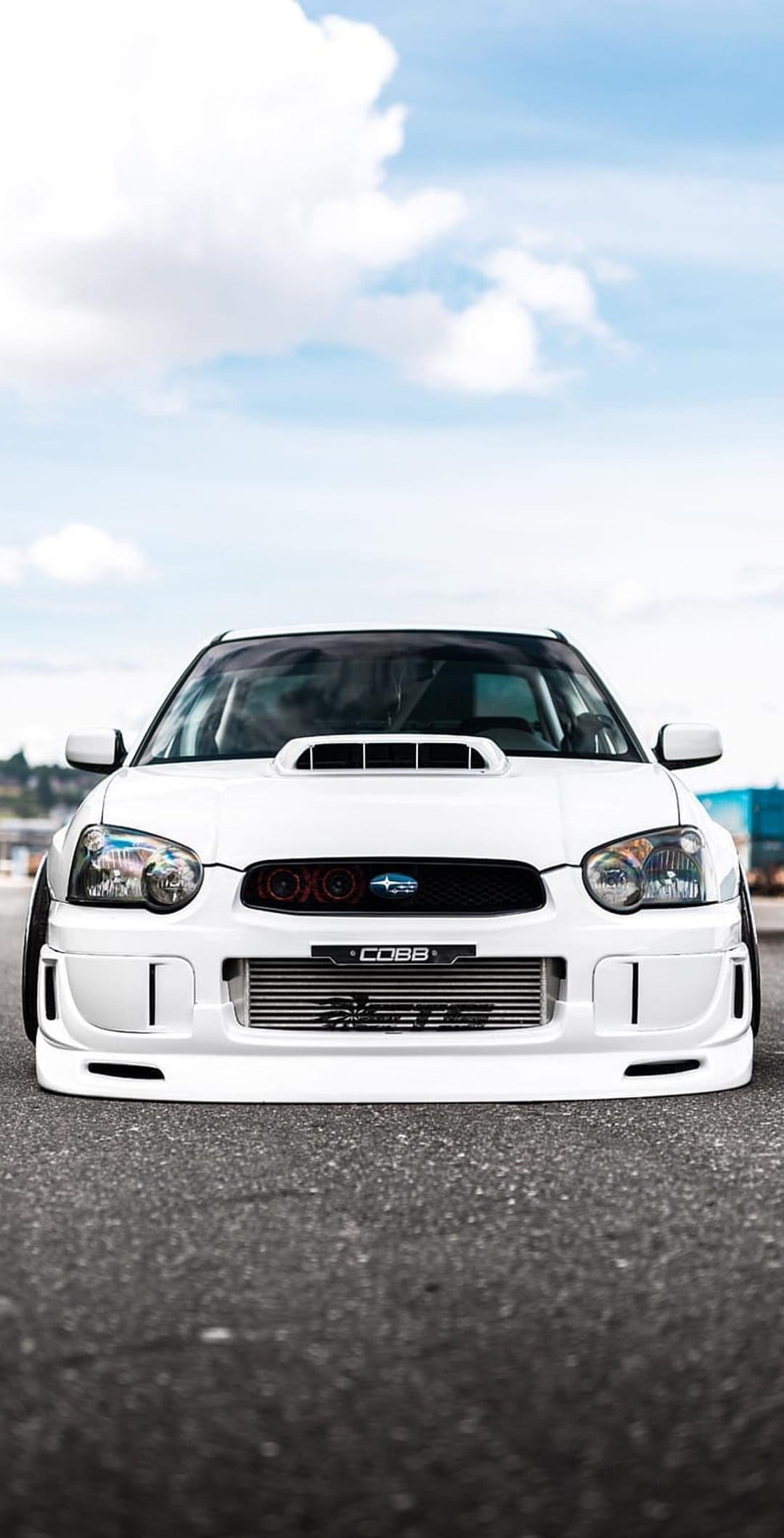 White Subaru WRX STI with a COBB intercooler parked on asphalt under a bright blue sky with white clouds.