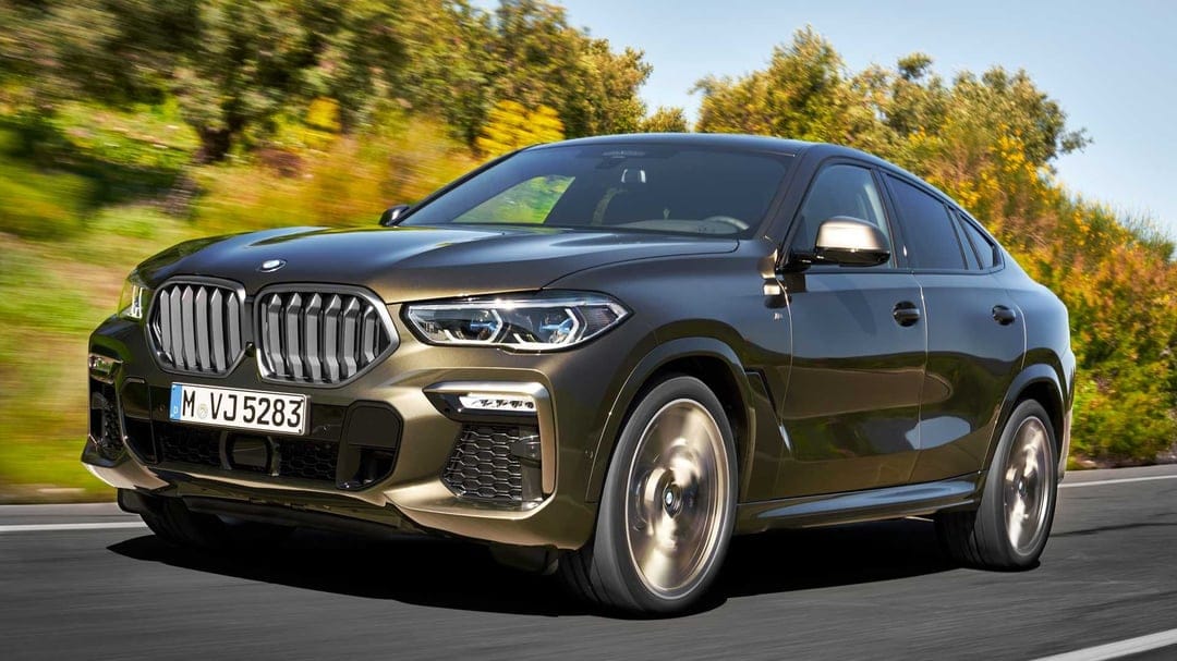 Bronze BMW X6 SUV driving on a sunny road with motion blurred wheels against a lush green forest background.