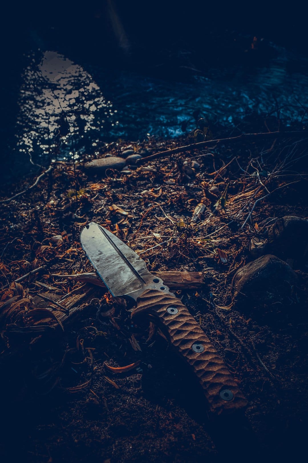 Tactical knife with a textured handle rests on a bed of damp autumn leaves beside a dark flowing forest stream.