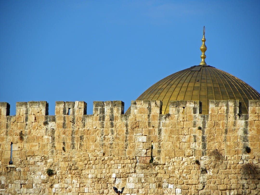 Golden Dome of the Rock rising behind a weathered stone wall under a clear blue sky in Jerusalem's Old City.