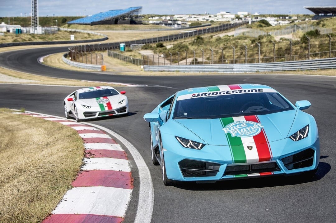 A light blue and a white Lamborghini Huracan racing on an asphalt track with red and white curbs and green grass.