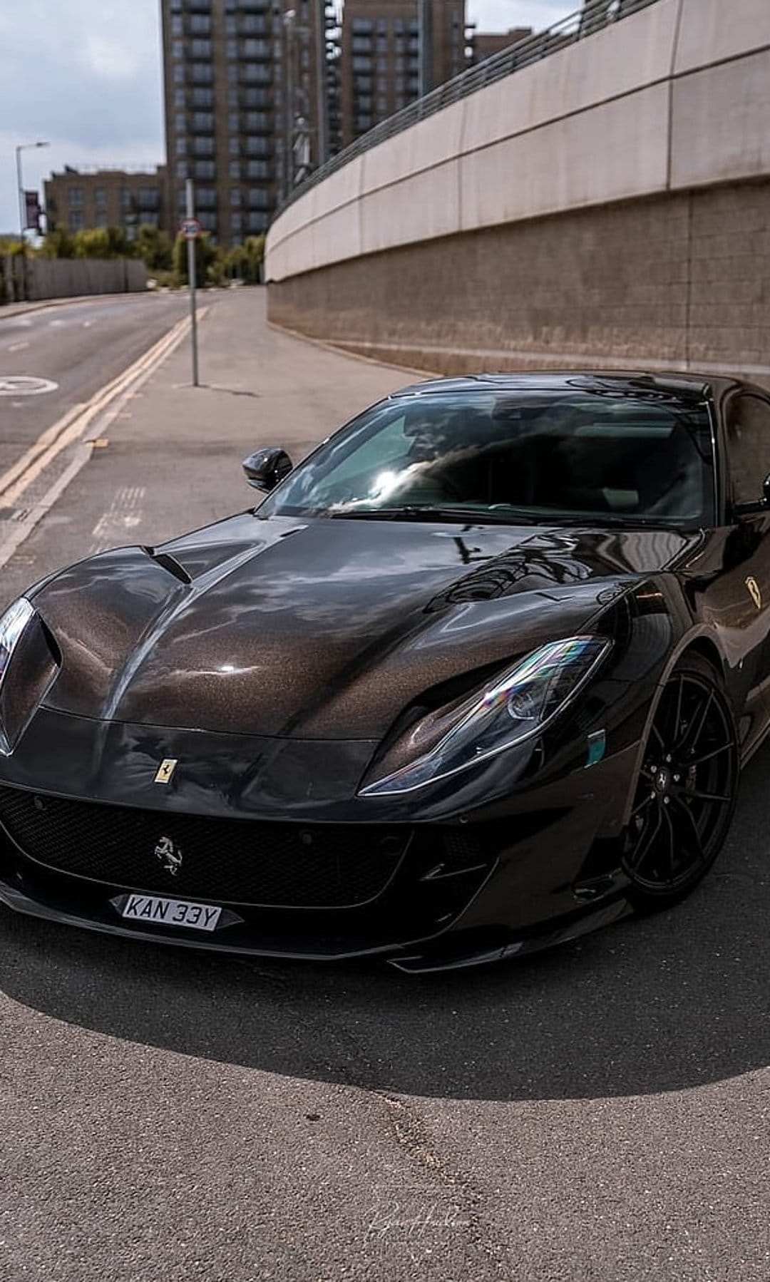 Ferrari 812 Superfast with dark metallic paint parked on an urban street near a curved wall at low angle.
