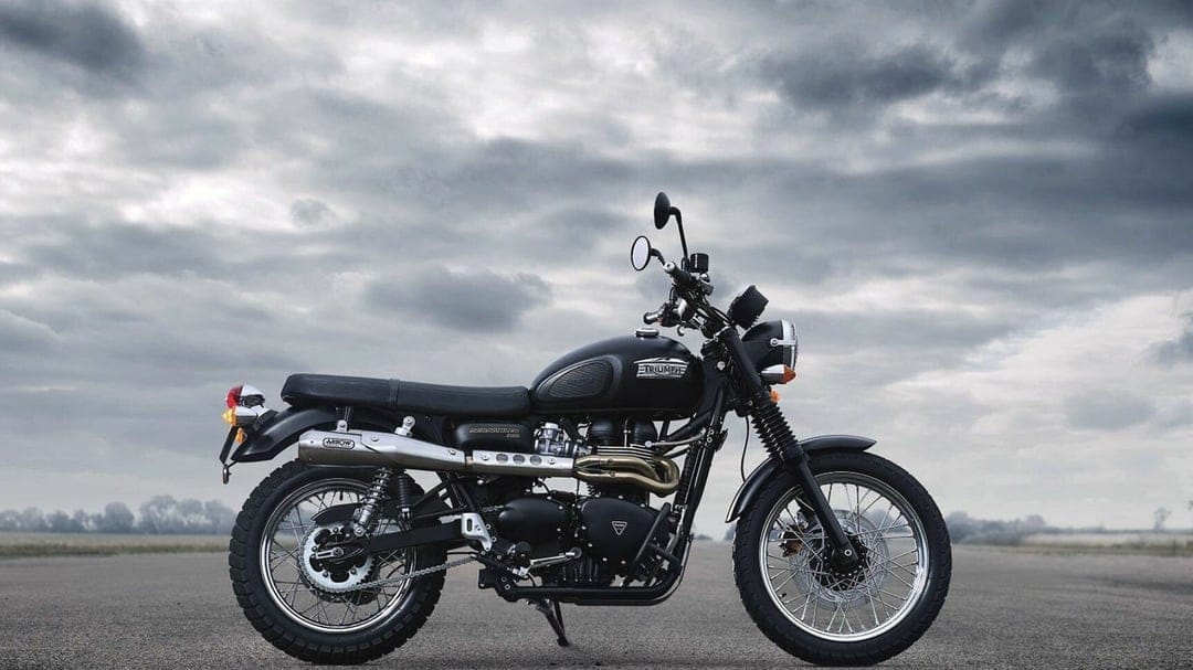 Profile of a black Triumph Scrambler motorcycle parked on open asphalt under a moody and dramatic overcast sky.