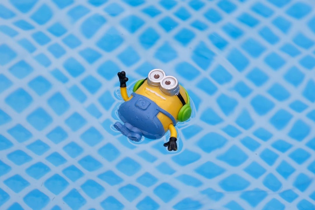 Minion toy with green headphones floats on a diamond-textured blue pool surface in a bright summer setting.
