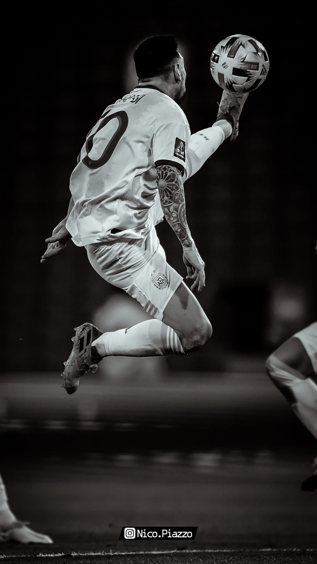 Lionel Messi leaps mid-air to control a football, displaying arm tattoos in a high-contrast monochrome scene.