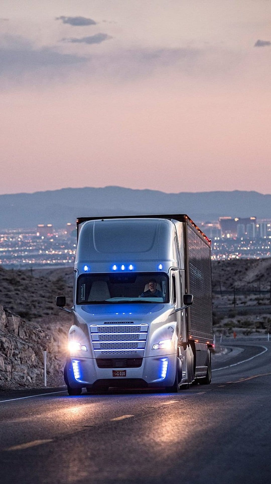 Sleek semi-truck with blue LED trim driving on a winding desert highway toward a glowing city at twilight.
