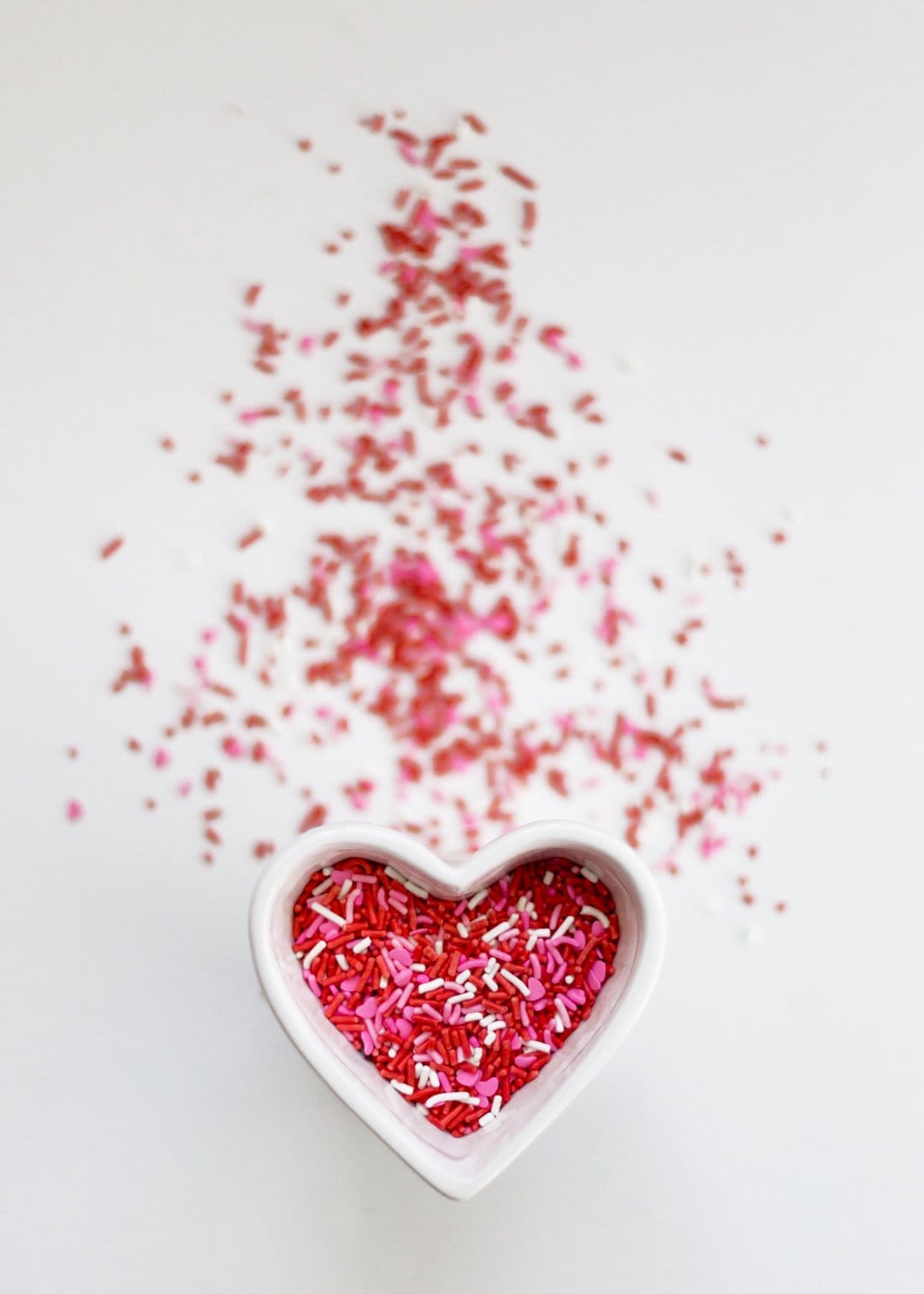 White heart-shaped bowl overflowing with red, pink, and white sugar sprinkles scattered on a bright white surface.