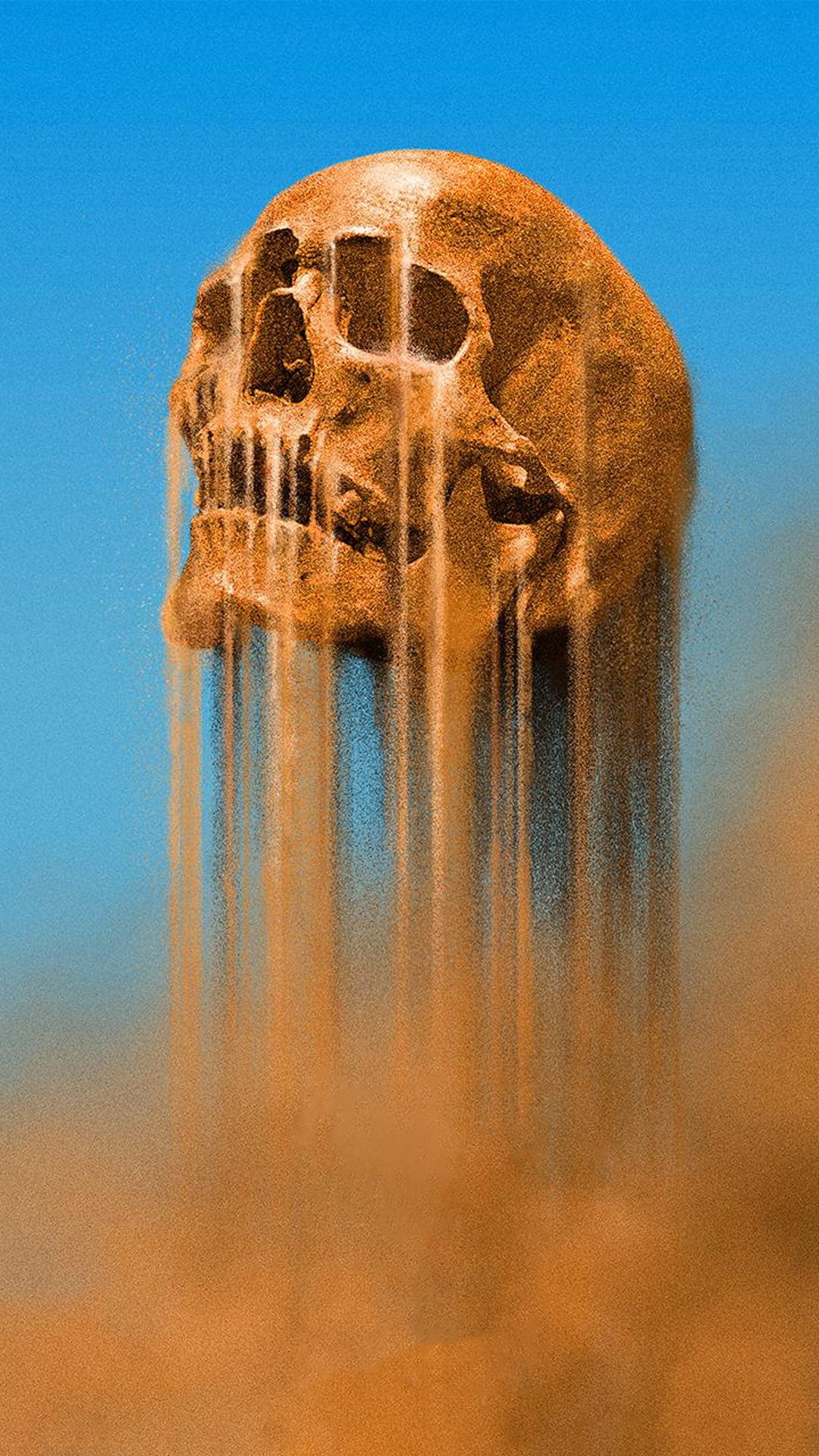 Orange skull fragmenting into fine sand streams against a solid blue sky with detailed bone and grit textures.