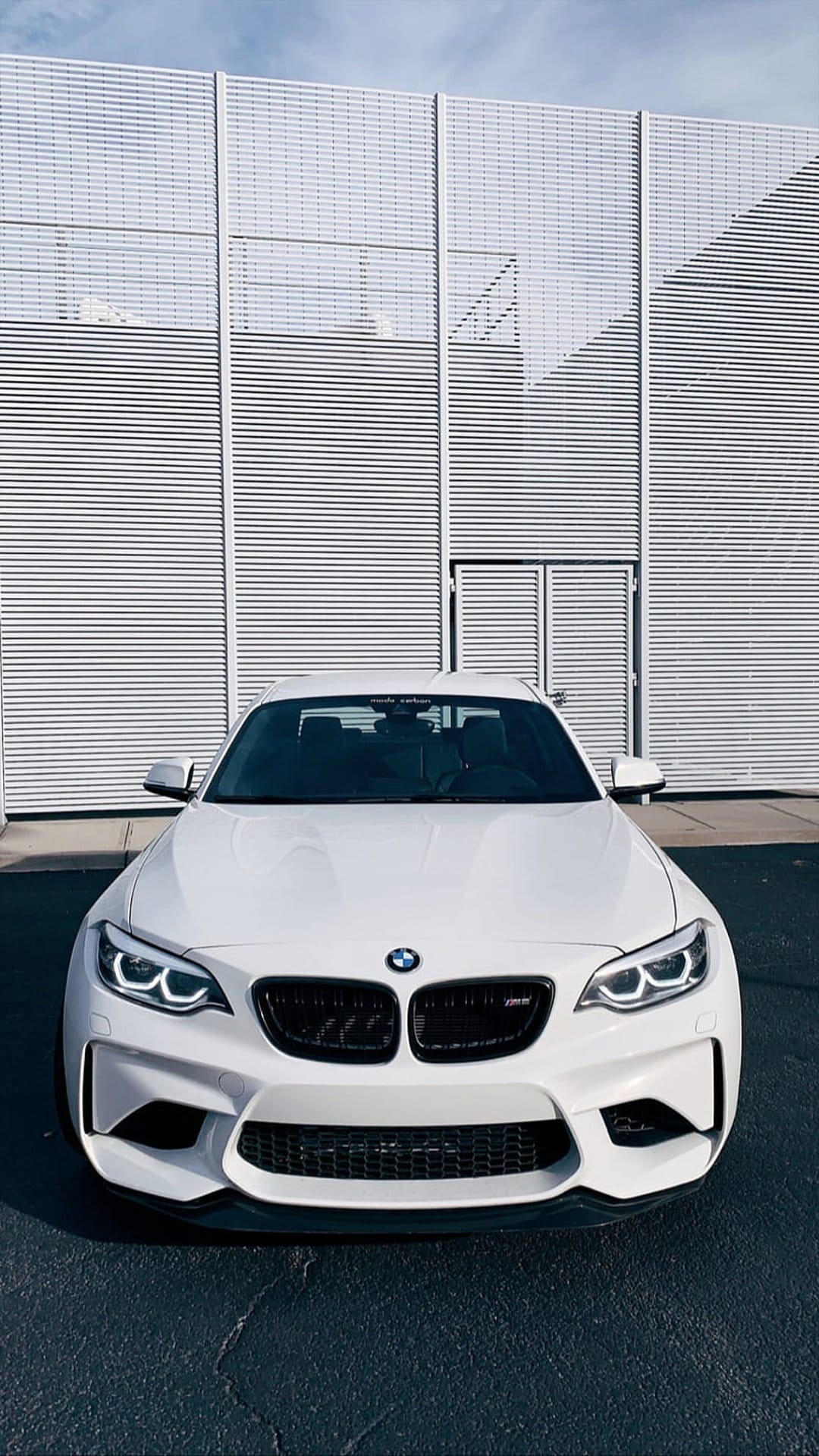 White BMW M2 sports car parked on asphalt in front of a modern building with metallic horizontal blinds and mesh.