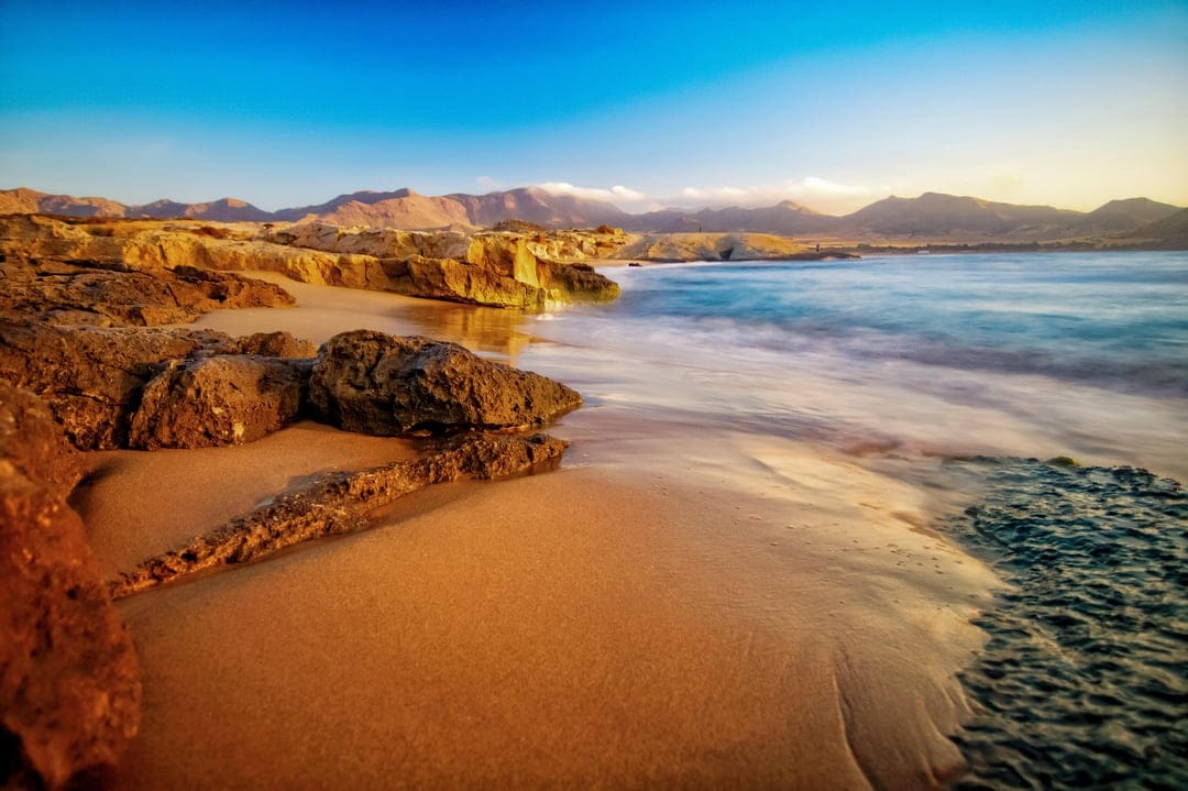 Rugged coastal rocks and misty blue ocean waves sit beneath a vibrant orange sunset and distant mountain peaks.