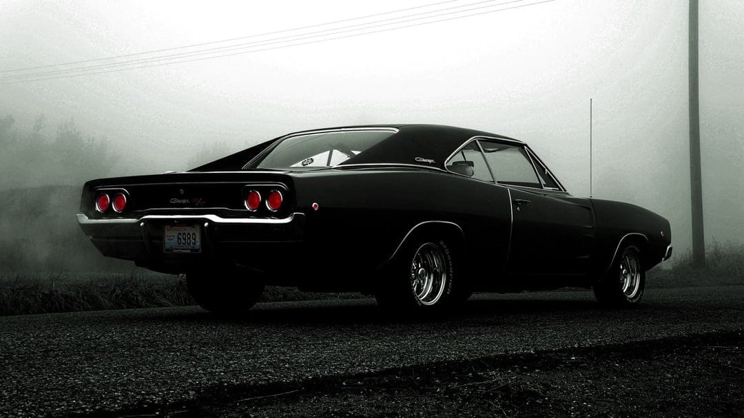 Black classic muscle car with glowing red tail lights parked on a misty road during a foggy day.