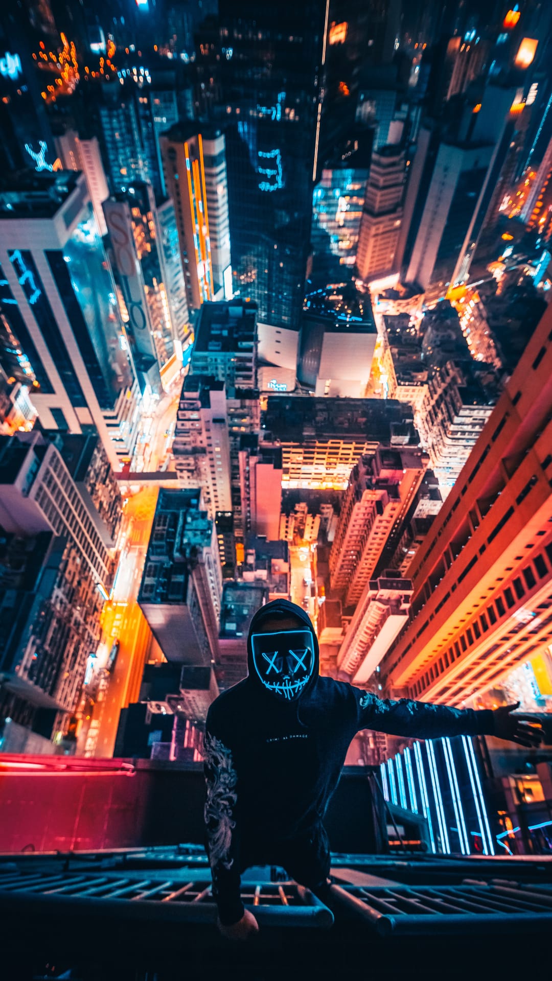 Masked person with glowing XX eyes kneels on a rooftop edge overlooking a vast, illuminated night cityscape.