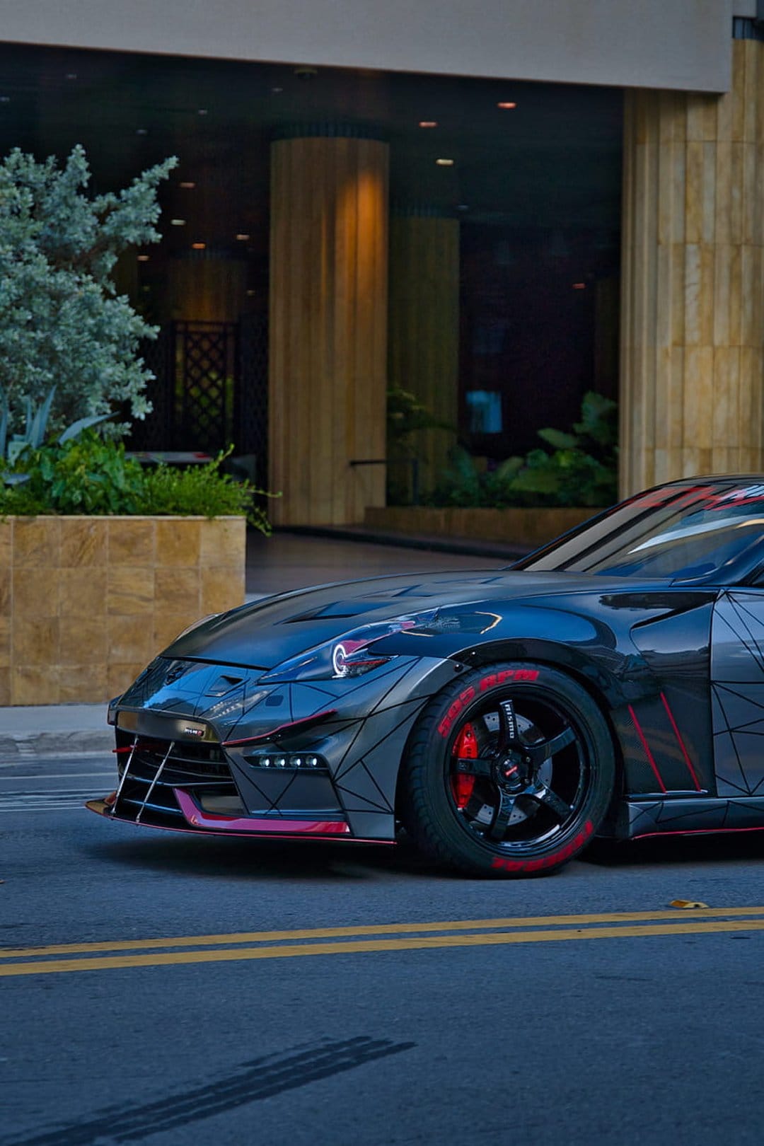 Custom sports car with red accents and a wide body kit parked on a city street beside modern glass architecture.
