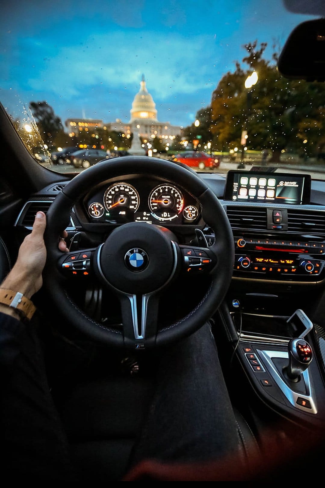 BMW steering wheel and dashboard interior facing the glowing U.S. Capitol Building through a rainy windshield.