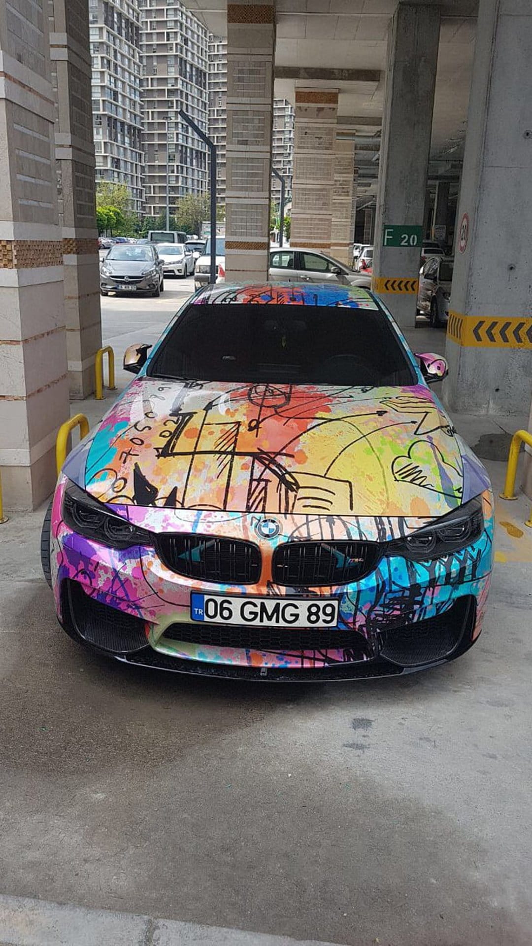 BMW M4 sports car with abstract graffiti wrap parked under a concrete structure in a modern city setting.