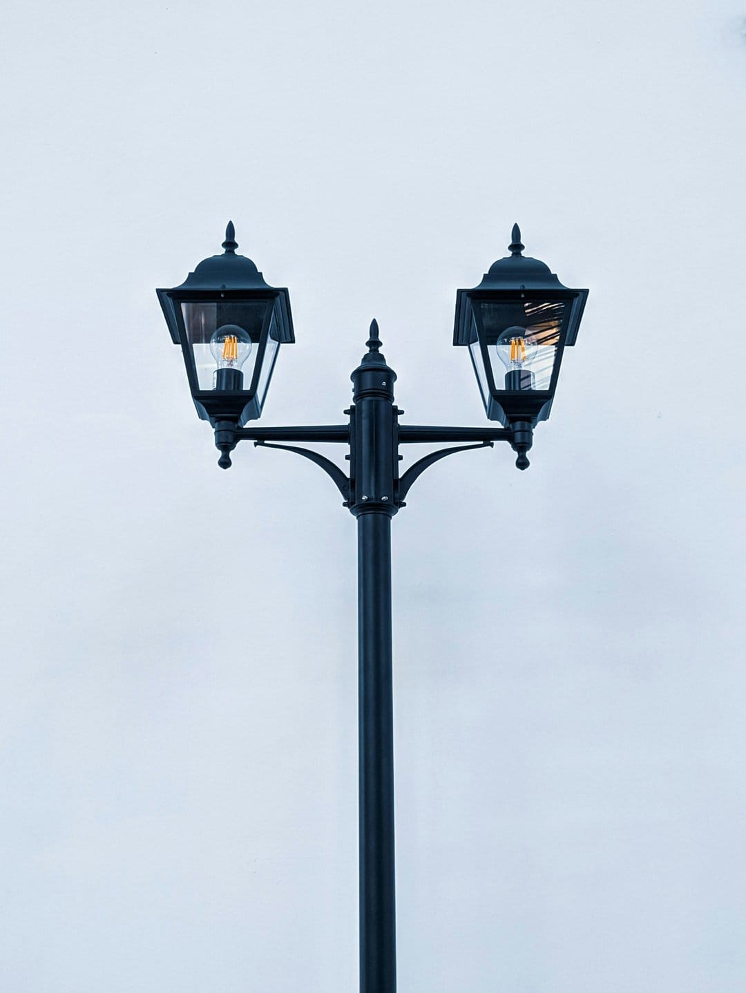 Black double lamp post with glowing bulbs centered against a pale blue sky in a vertical composition.