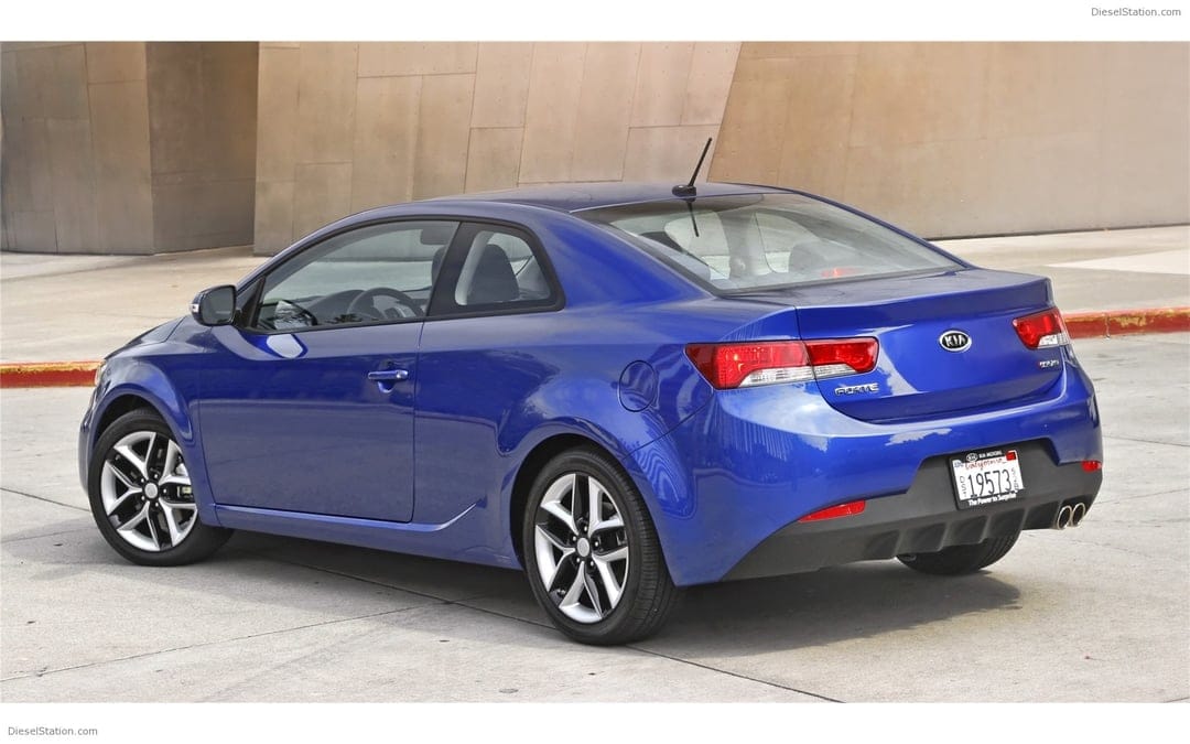 Blue Kia Forte Koup sports coupe parked on concrete with a modern metallic architectural wall in the background.
