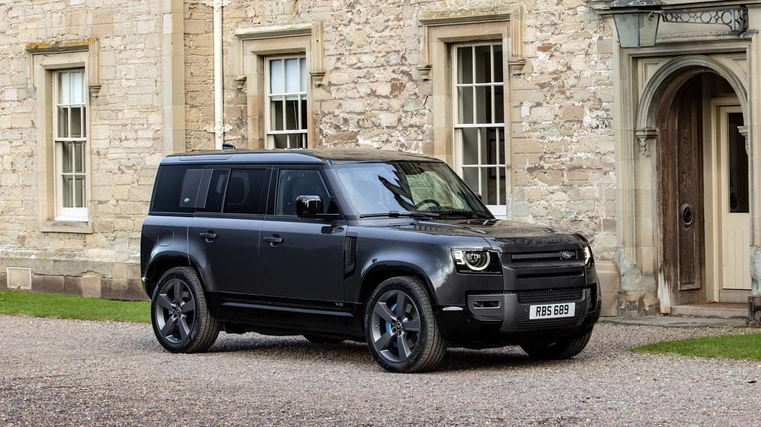 Dark grey Land Rover Defender V8 parked on gravel in front of a historic stone manor house with large windows.