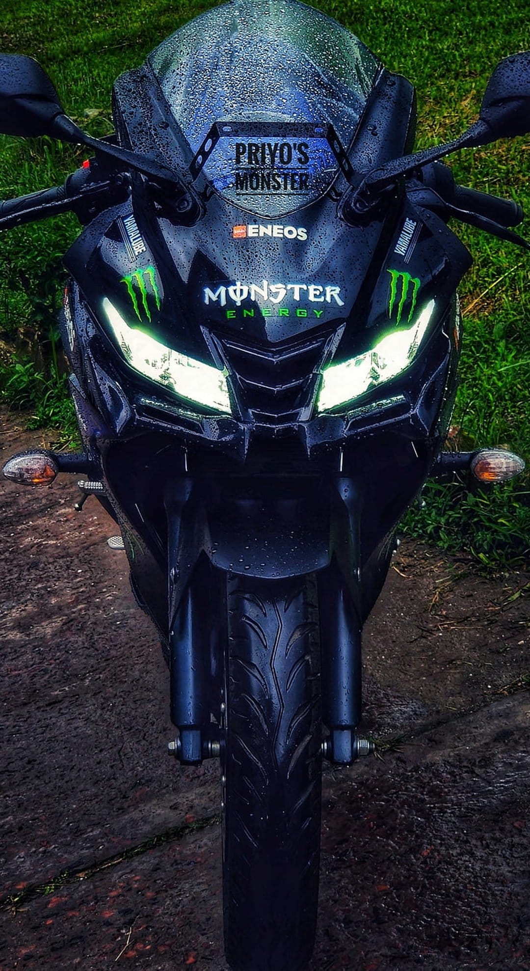 Yamaha R15 motorcycle with glowing headlights and Monster Energy decals parked on wet grass during a rainy day.