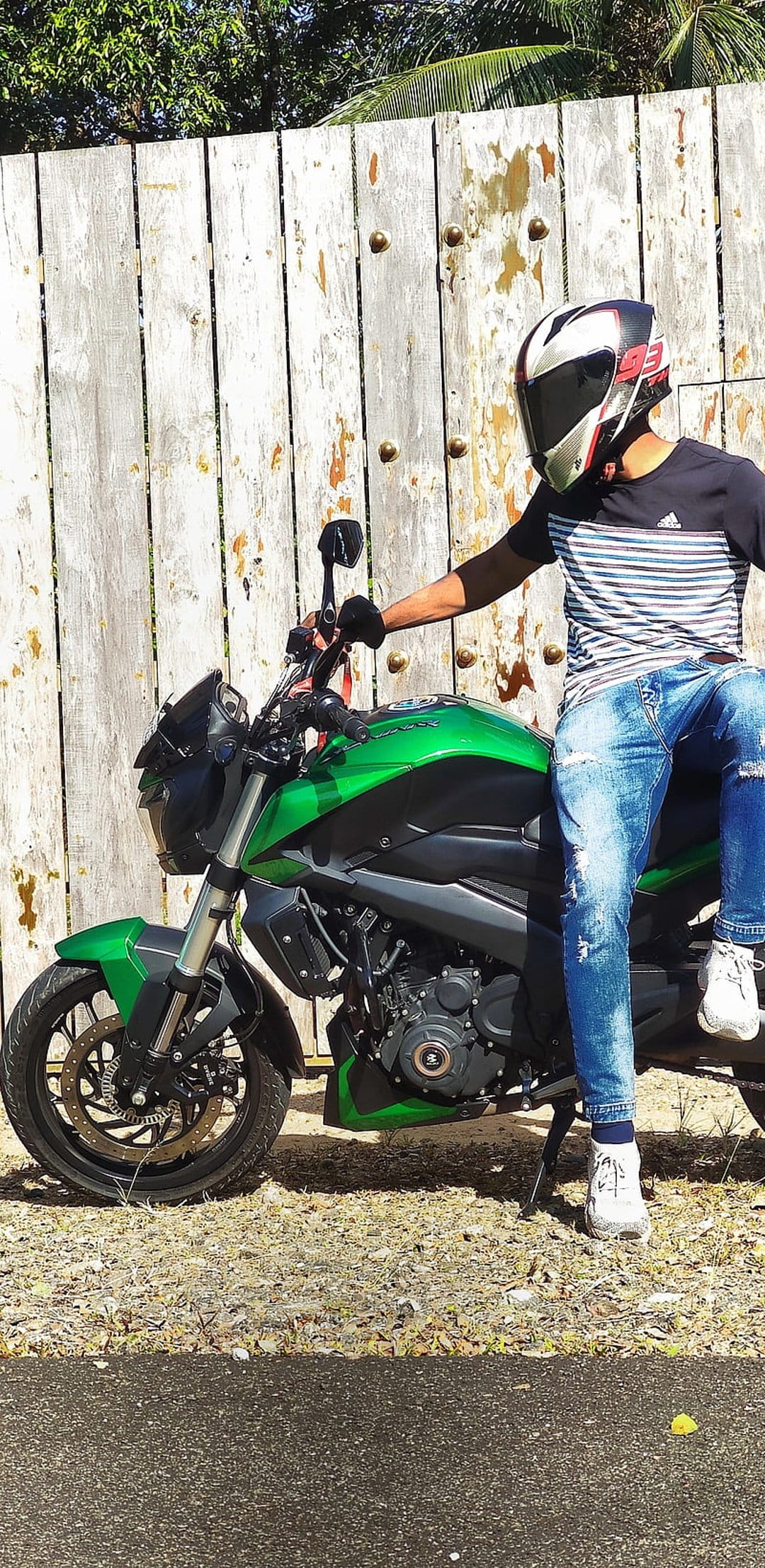 Motorcyclist in a helmet and jeans sits on a lime green bike beside a weathered wooden fence in the sun.