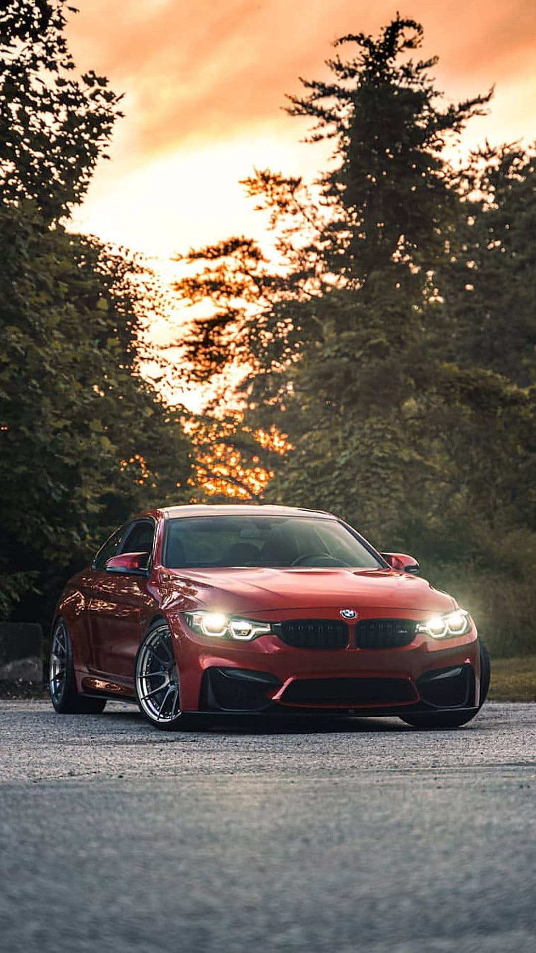 Red BMW M4 Coupe with glowing headlights parked on a road amidst green trees during a vibrant orange sunset.
