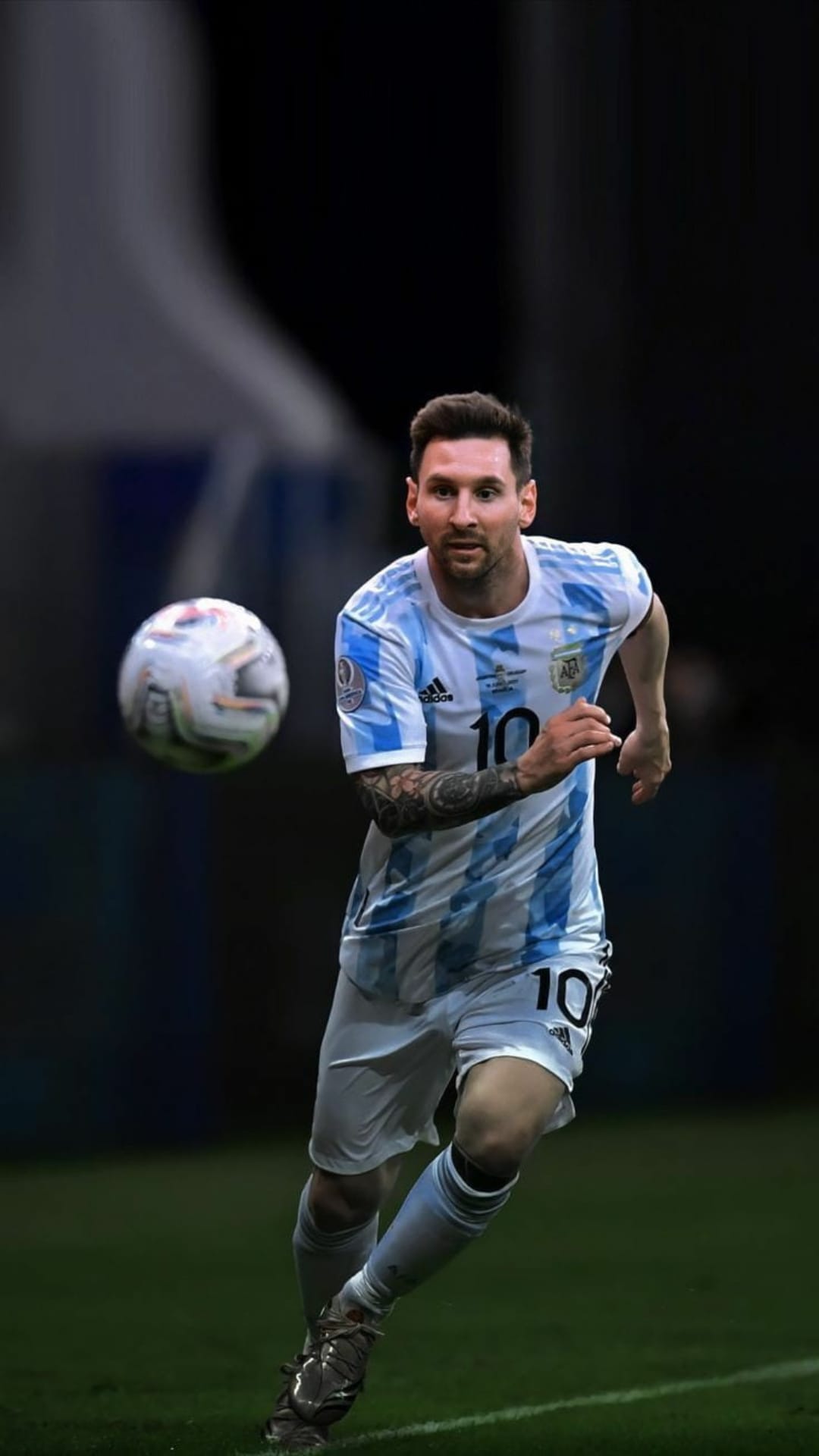 Lionel Messi sprints toward a soccer ball in an Argentina jersey on a dark green field with stadium lighting.