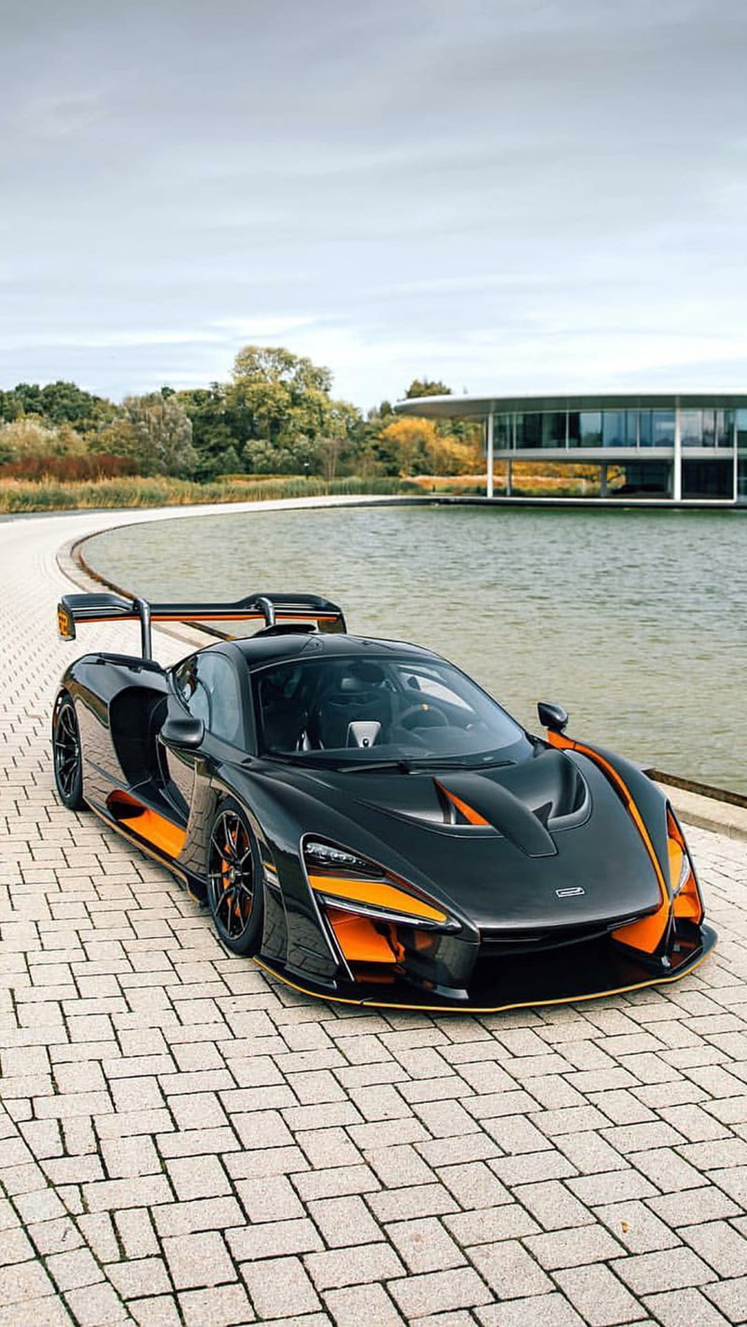 Black McLaren Senna supercar with orange accents parked on cobblestones near a glass building and waterfront.