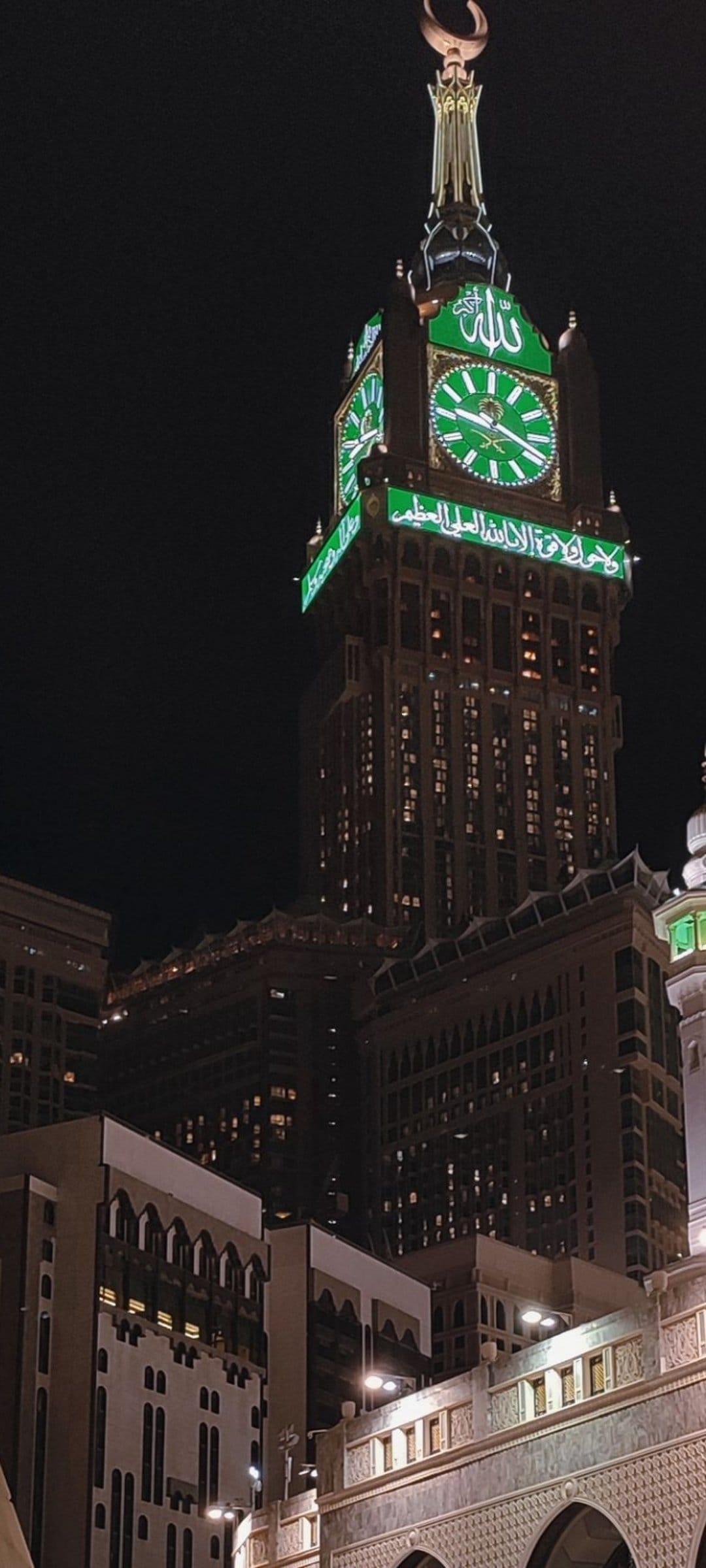 Makkah Clock Tower glows with green lights and Arabic calligraphy under a crescent moon against a dark night sky.