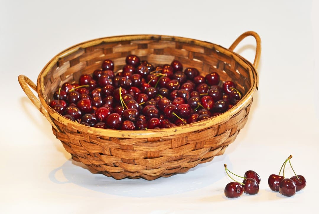 Ripe dark red cherries with green stems spill out of a rustic wicker basket onto a flat, white surface.