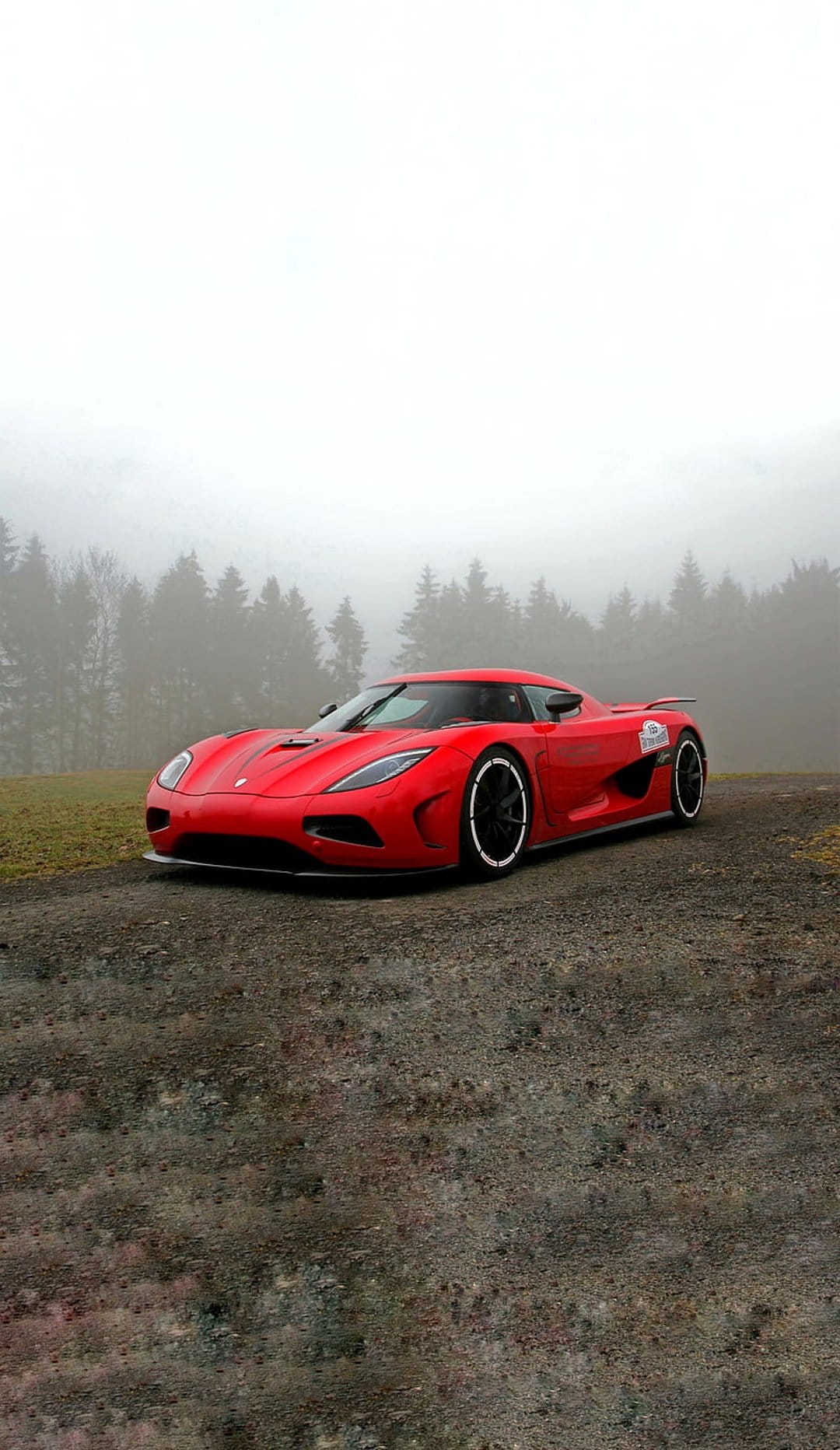 Red Koenigsegg supercar on a gravel road surrounded by a dense, foggy forest with tall trees in soft lighting.