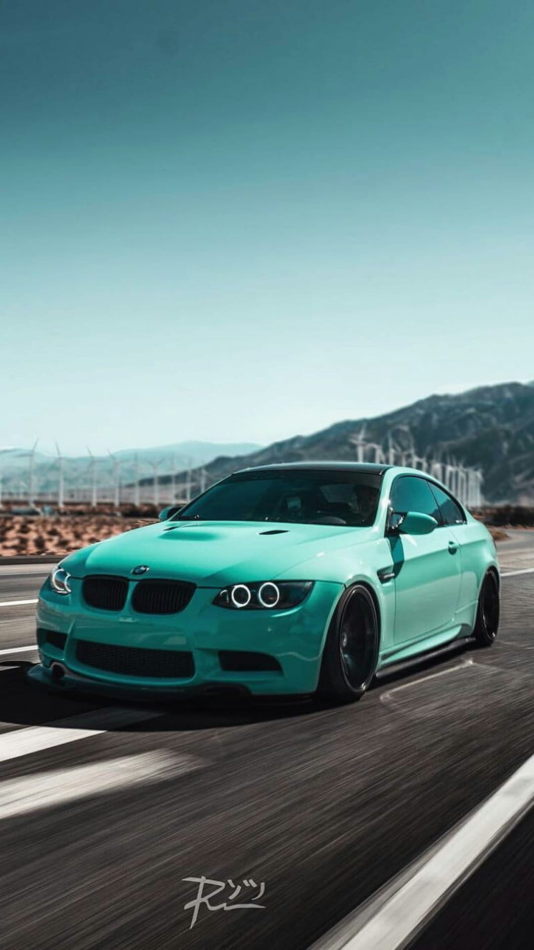 Teal BMW M3 coupe with black rims driving on a desert road featuring wind turbines and distant mountain peaks.