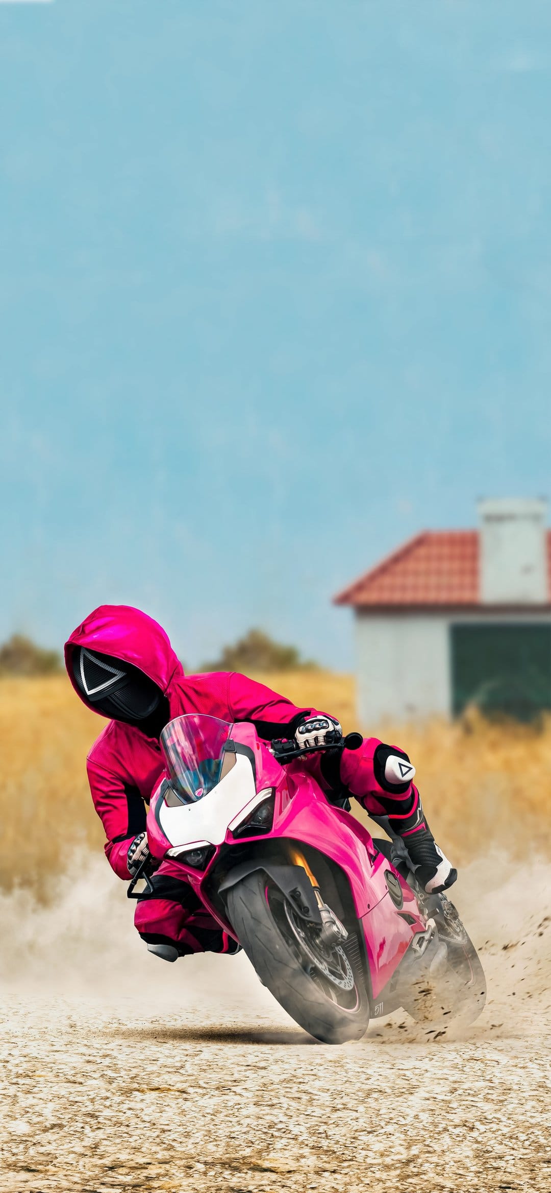 Masked Squid Game guard drifting a pink motorcycle on a dirt road near a red-roofed house in a golden field.