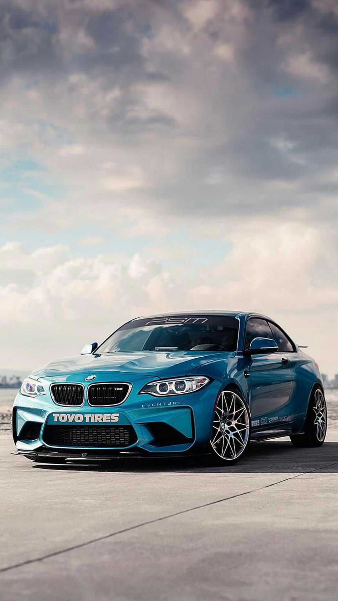 Blue BMW M2 CS sports car with Toyo tires on a paved road against a blurred ocean and cloudy sky background.
