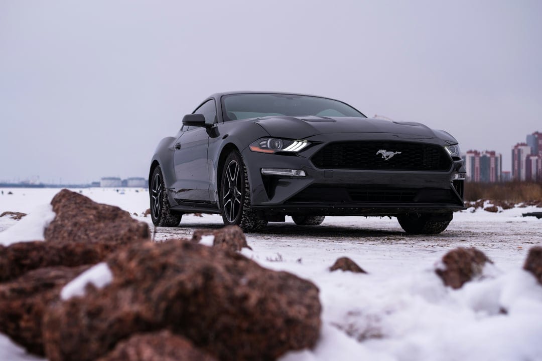 Dark Ford Mustang parked on snowy ground with foreground rocks and a distant city skyline under cloudy skies.
