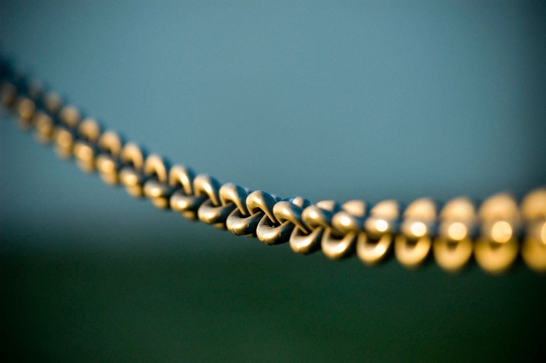 Golden metal chain links shimmer under warm sunlight against a soft blue and green blurred background.
