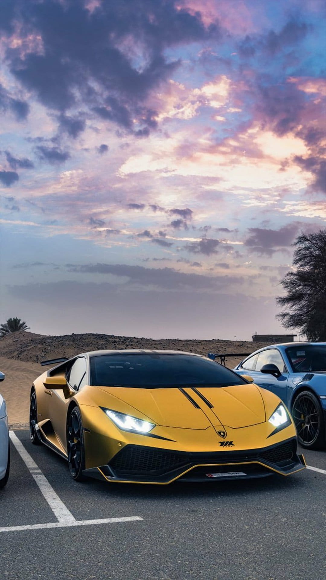 Golden Lamborghini and blue Porsche parked on desert sand beneath a vibrant orange and purple sunset sky.