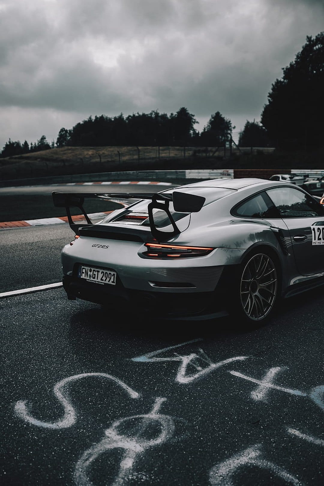 Silver Porsche 911 GT3 RS with glowing red taillights on a wet race track under a dark, moody and cloudy sky.