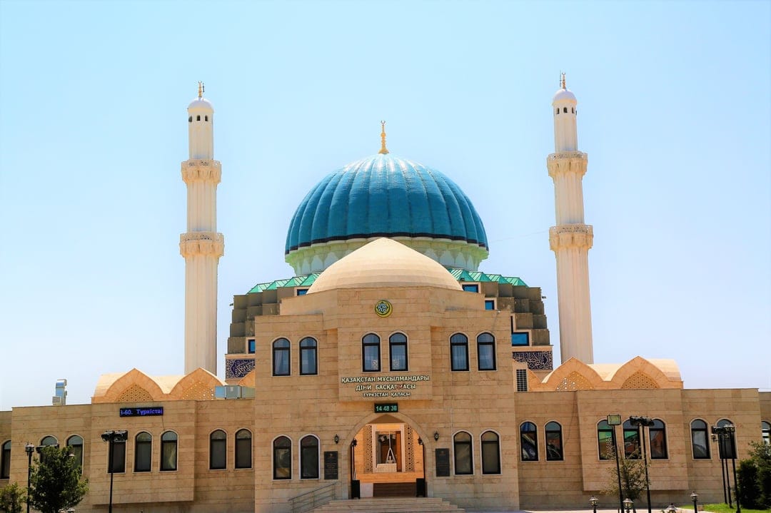 Ornate blue dome mosque with twin minarets and arched windows stands against a clear, sunny sky in midday light.